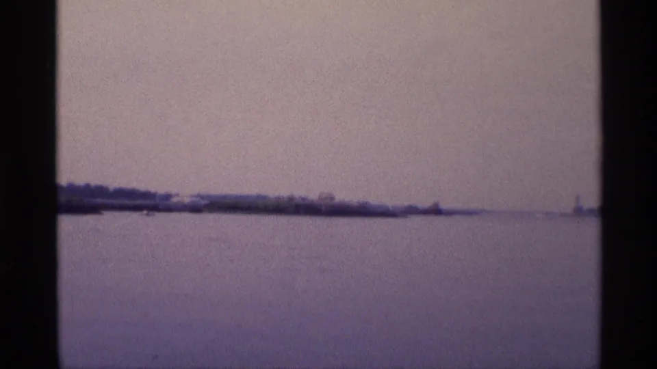 Faded 1973 New York 8mm frame with calm water, low shoreline, and a small distant vertical structure near the horizon.