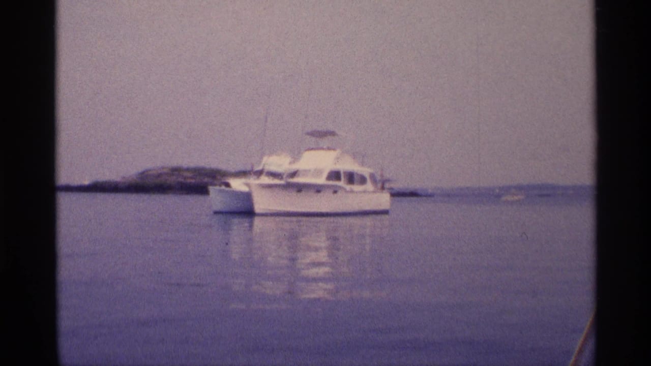1973 New York 8mm frame showing a white motorboat on calm water near a low rocky outcrop and distant shore.
