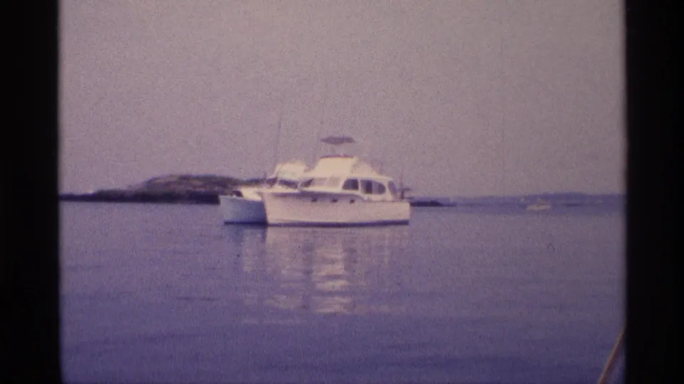 1973 New York 8mm frame showing a white motorboat on calm water near a low rocky outcrop and distant shore.