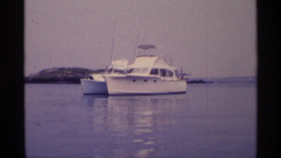 Soft 8mm frame of a white motorboat in calm New York-area water, with a low shoreline behind it.