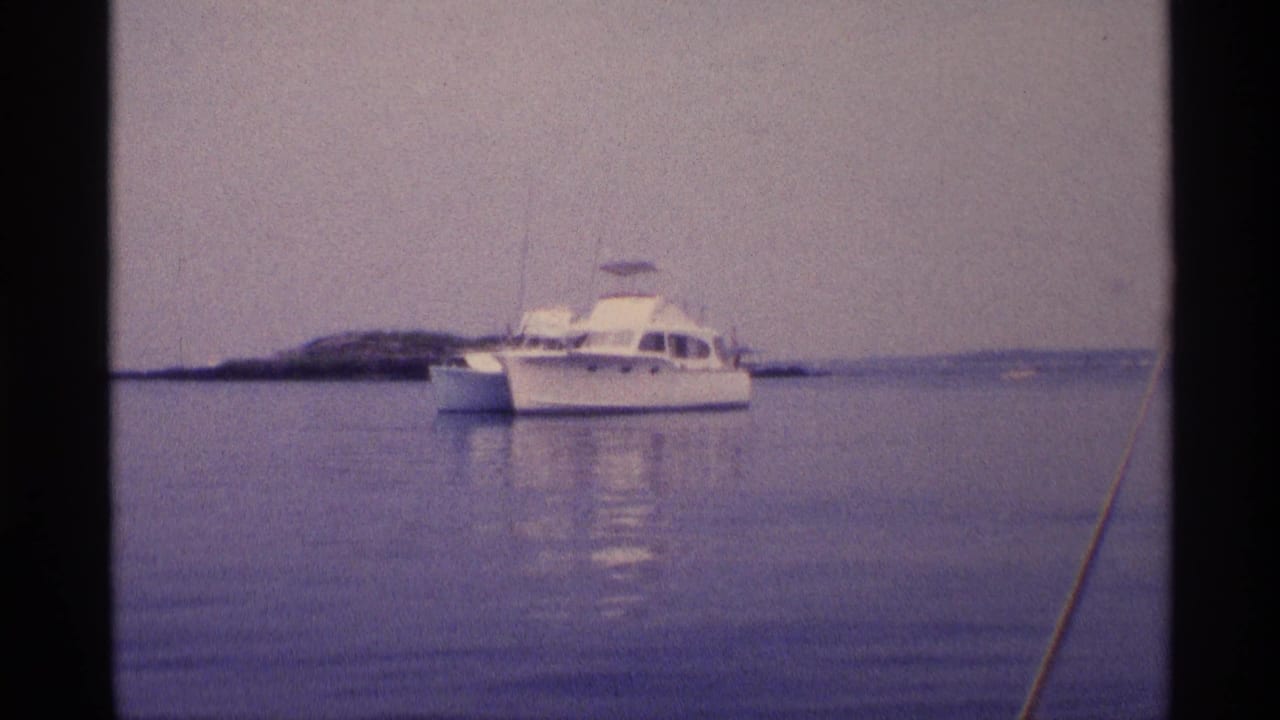 Wide 1973 New York 8mm view of a white motorboat at anchor on still water near a low island or shoreline.