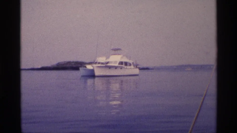 Wide 1973 New York 8mm view of a white motorboat at anchor on still water near a low island or shoreline.