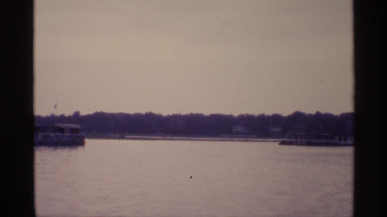 Distant boats pass across calm New York lake water in a soft, grainy 1973 home-movie frame.
