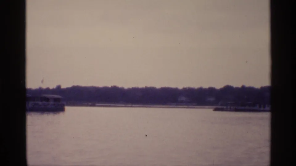 Distant boats pass across calm New York lake water in a soft, grainy 1973 home-movie frame.
