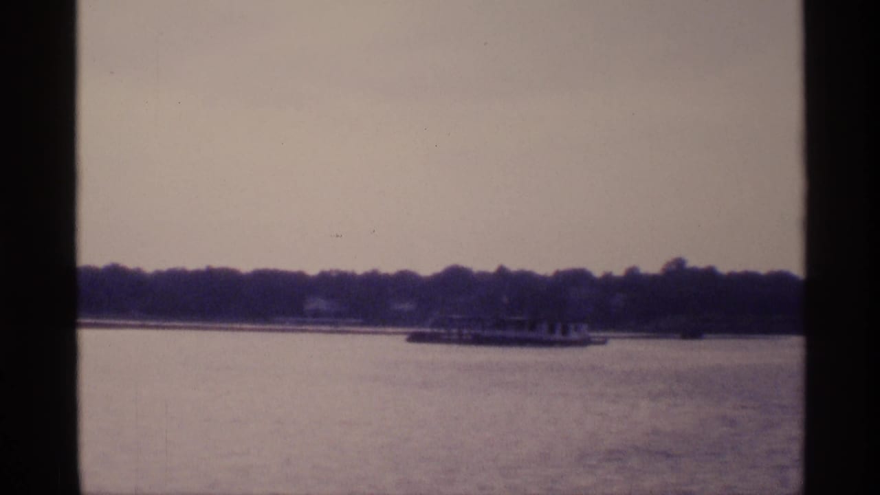 A distant boat moves along calm New York lake water, framed by wooded shoreline and aged 8mm texture.