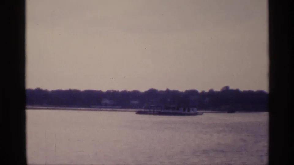 A distant boat moves along calm New York lake water, framed by wooded shoreline and aged 8mm texture.