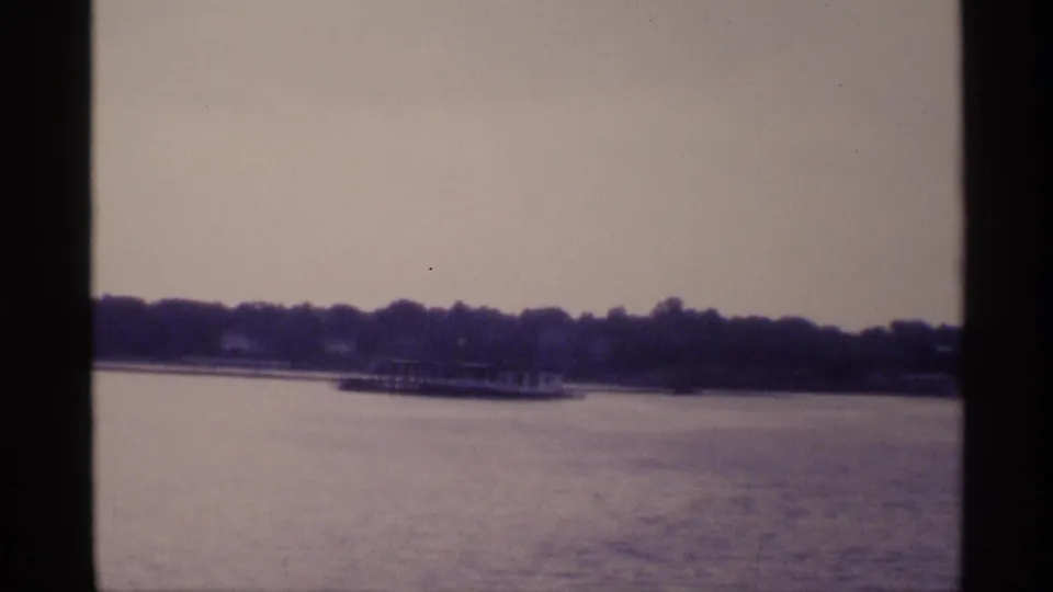 1973 New York 8mm view from a boat toward calm water, a tree-lined shore, and low distant lakeside structures.