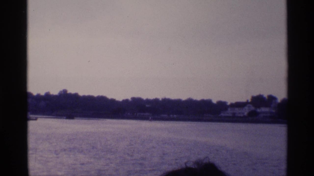 From a moving boat, the 1973 New York home movie looks toward a wooded shore with a small structure and partial passenger visible.