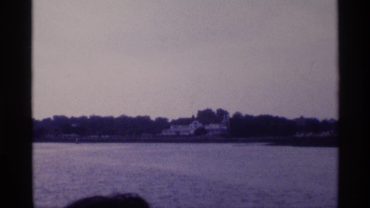 The pan settles on a distant white lakeside house among trees across the water, with a partial boat passenger visible in the foreground.