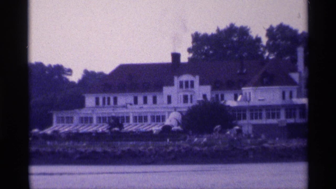 Closer 8mm frame of the white riverside house, showing porch windows, roof details, trees, and rocky shoreline.