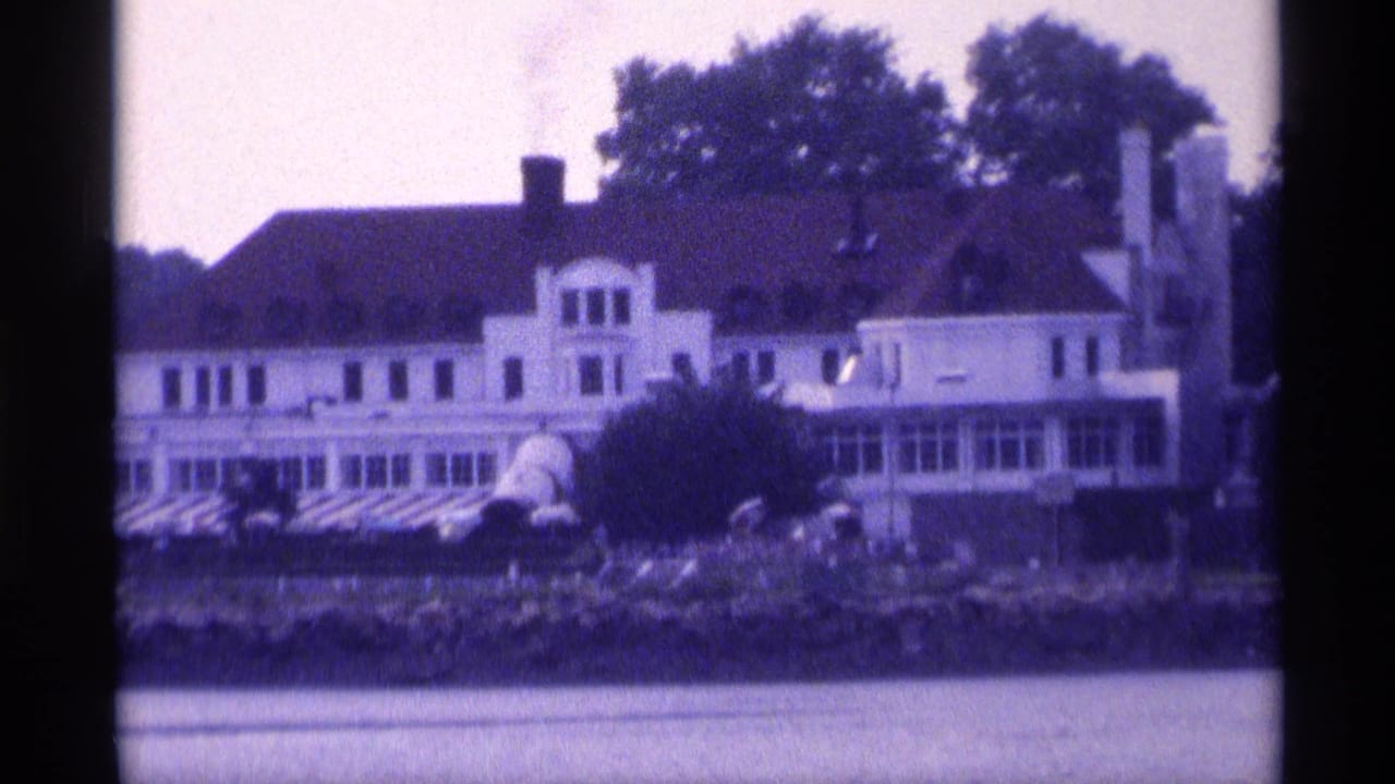 Close 1973 New York home-movie view of the large white house beside the water, with chimneys, porch glazing, shrubs, and shoreline rocks.