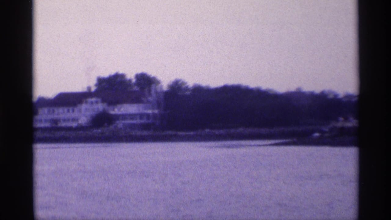 Distant 1973 New York waterfront view of a white house along a wooded shoreline, filmed across a broad stretch of calm water.