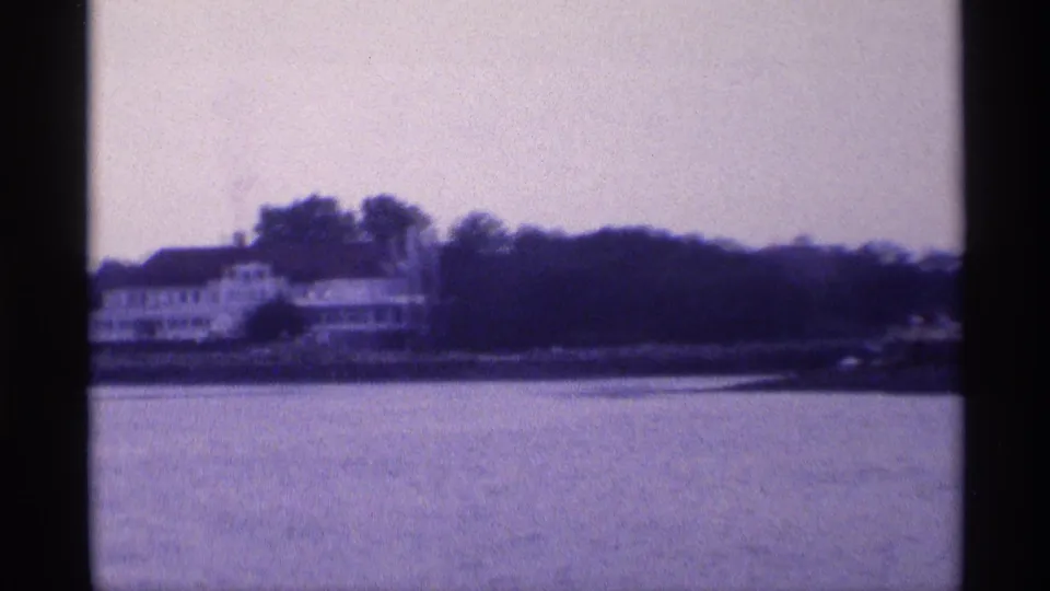 Distant 1973 New York waterfront view of a white house along a wooded shoreline, filmed across a broad stretch of calm water.