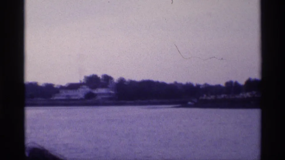 Wide 8mm frame from 1973 New York showing the white waterfront house farther away, with trees and calm water dominating the scene.