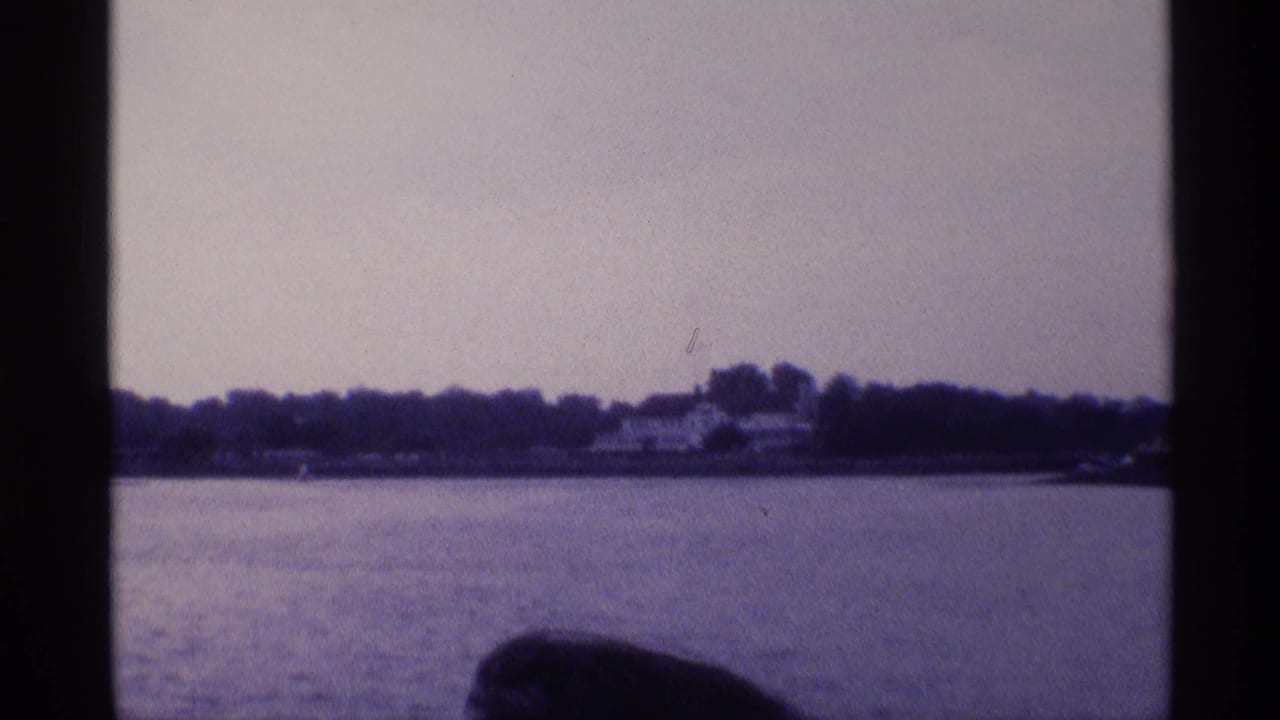 Final 1973 New York frame showing the waterfront house at a distance, with calm water in front and a dark foreground shape near the lower edge.
