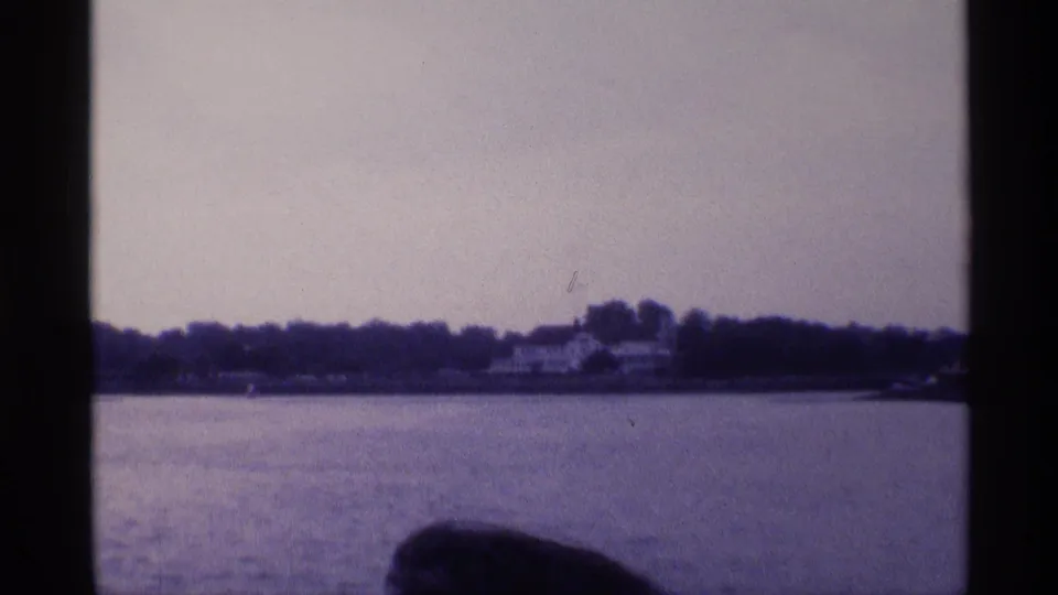 Final 1973 New York frame showing the waterfront house at a distance, with calm water in front and a dark foreground shape near the lower edge.
