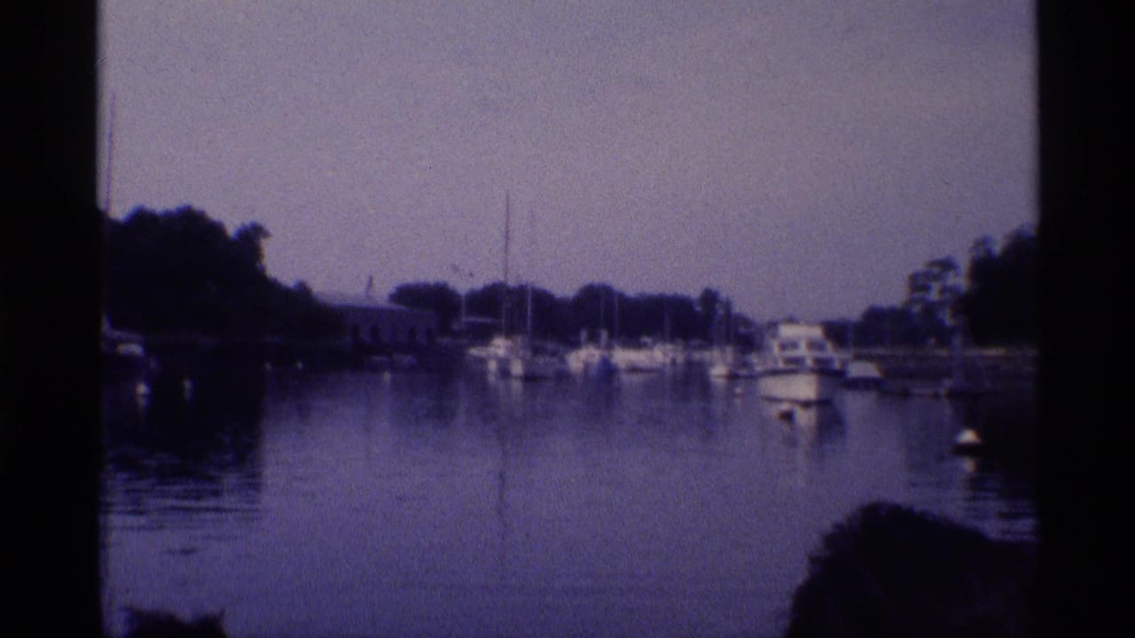 1973 New York waterfront frame showing docked sailboats, a small motorboat, calm water, trees, and shoreline structures.