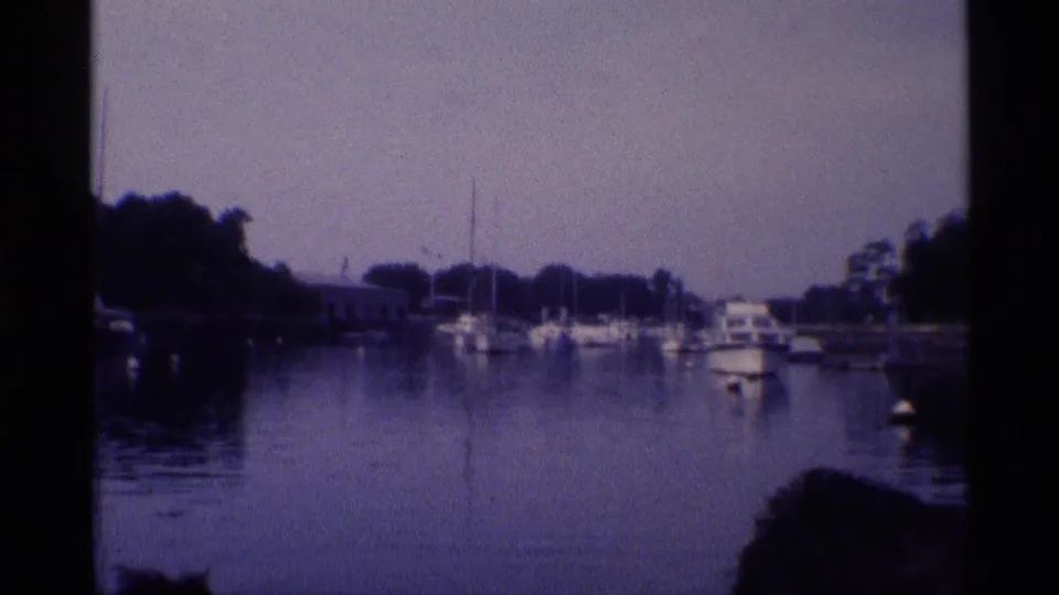 1973 New York waterfront frame showing docked sailboats, a small motorboat, calm water, trees, and shoreline structures.