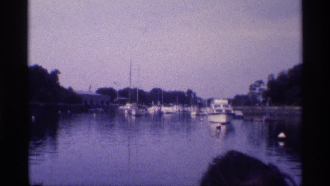 1973 New York 8mm frame of a marina with sailboat masts, a small motorboat, calm reflections, trees, and a foreground head silhouette.