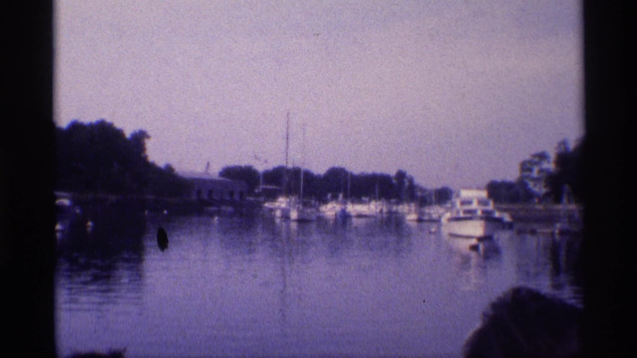 Aged 8mm frame from 1973 New York showing boats at a marina, calm water, wooded shoreline, and a partial foreground head silhouette.