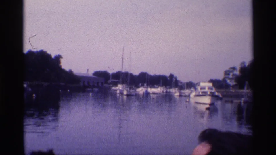 1973 New York 8mm frame showing a marina or canal with boats, shoreline trees, and a partial foreground spectator.