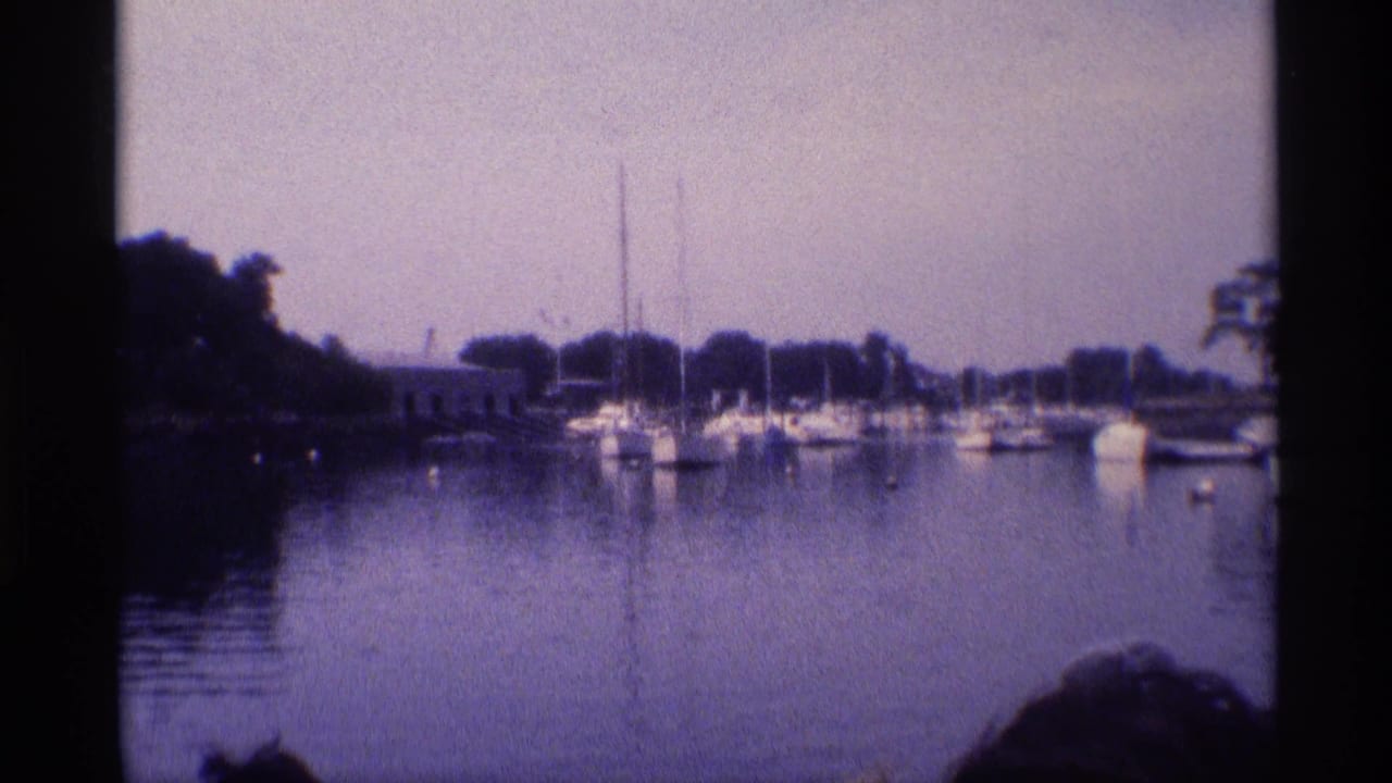 1973 New York home-movie frame of sailboats and small boats moored across still marina water under a hazy sky.