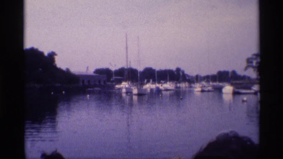 1973 New York home-movie frame of sailboats and small boats moored across still marina water under a hazy sky.