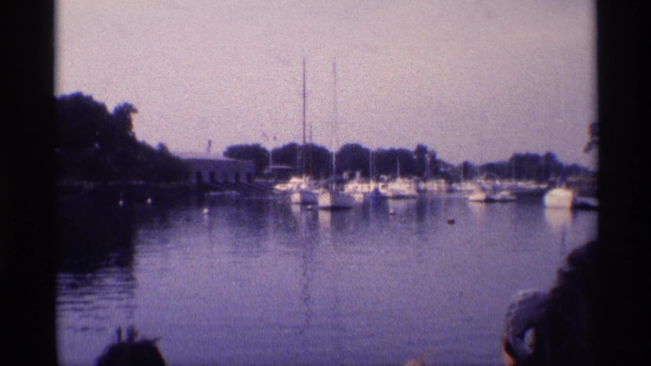 1973 New York home-movie frame showing a quiet marina scene with boats, masts, trees, and a partial foreground viewer.