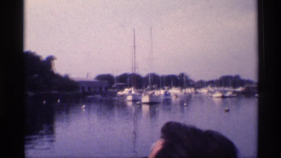 Wide 1973 New York 8mm view across a marina, with moored sailboats, dark trees, cloudy sky, and a foreground head.