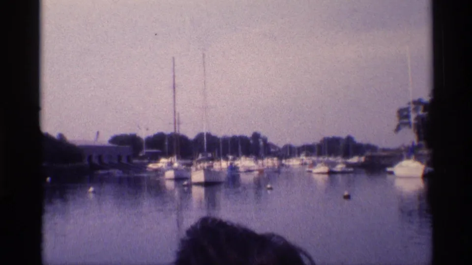 1973 New York 8mm frame showing moored sailboats across calm marina water, a tree-lined shore, and a partial foreground head.