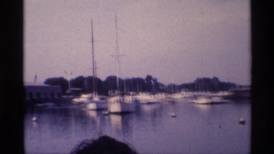 1973 New York 8mm view of a quiet marina with boats moored near a wooded shoreline and a partial head at the lower edge.