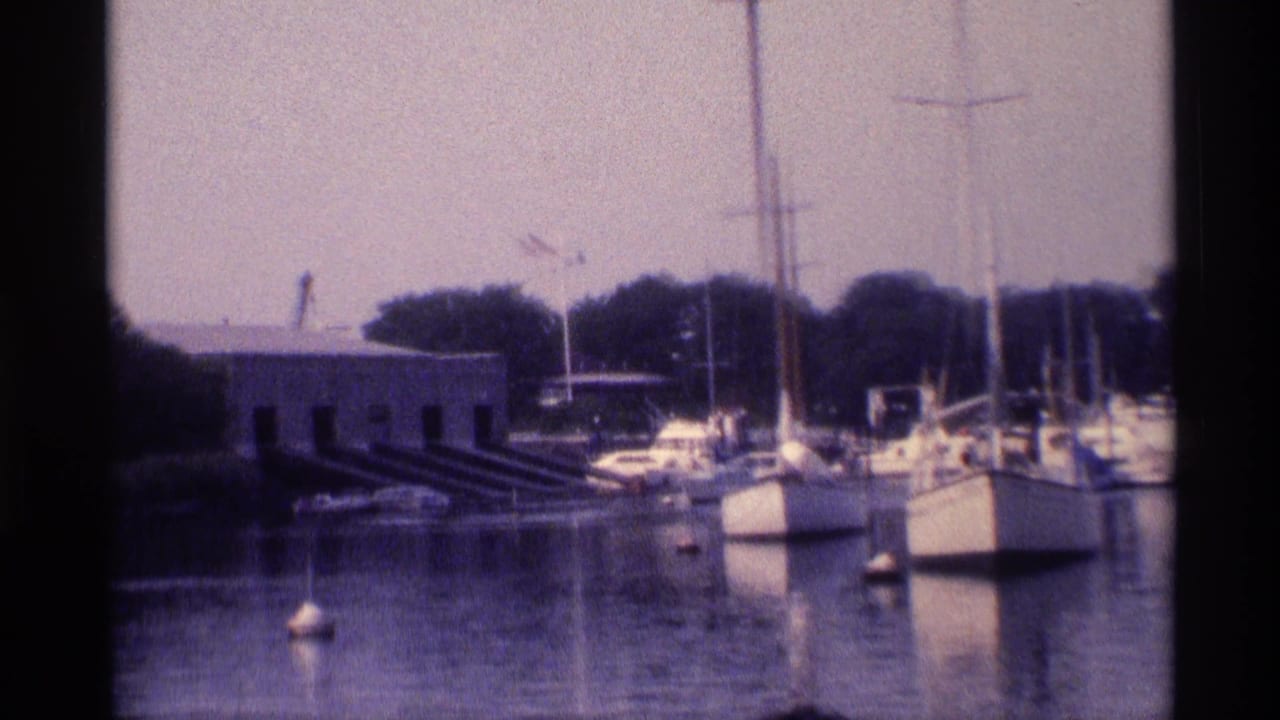 1973 New York home-movie frame showing sailboats and buoys near a low waterfront building with trees behind the marina.