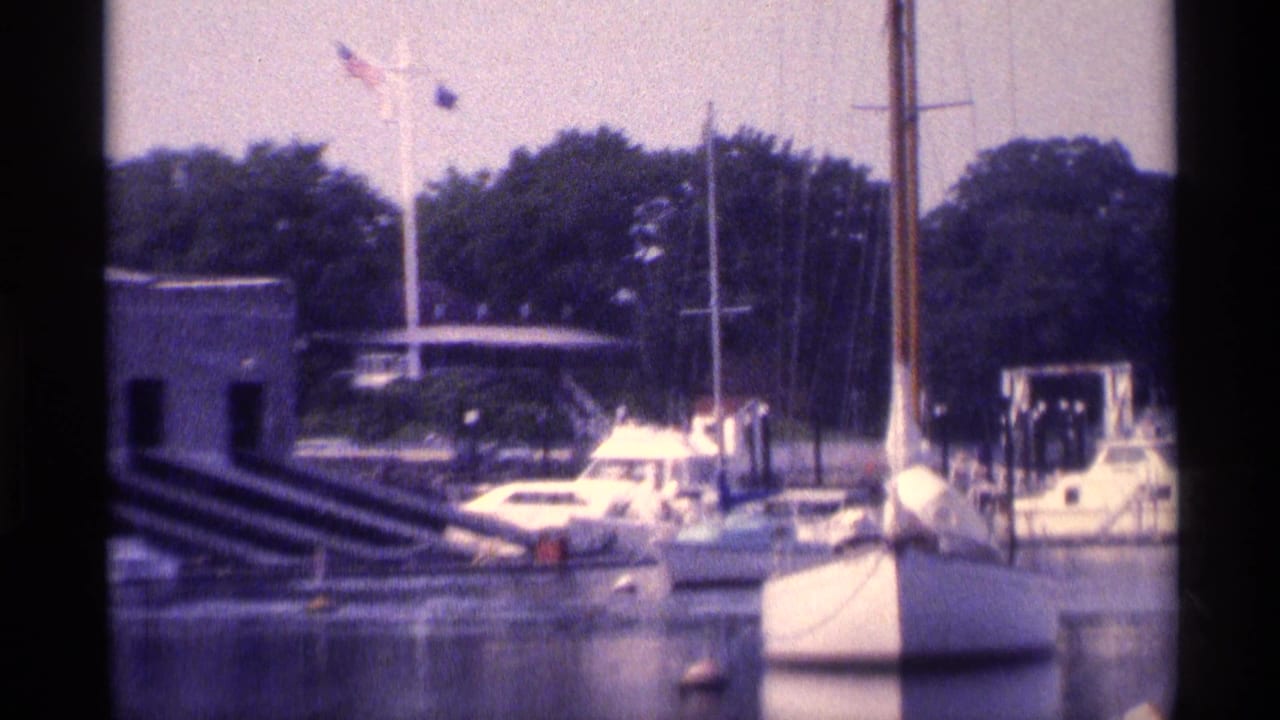 1973 New York 8mm frame showing a closer marina section with a white boat, masts, buoy markers, trees, and a low waterfront structure.
