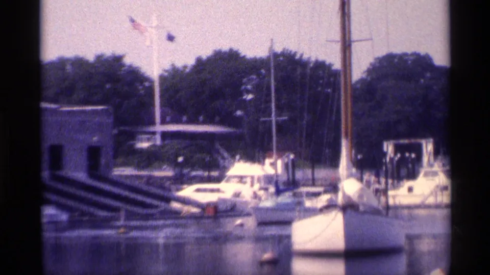 1973 New York 8mm frame showing a closer marina section with a white boat, masts, buoy markers, trees, and a low waterfront structure.