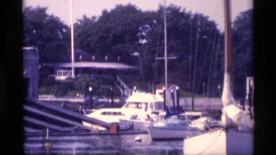 1973 New York 8mm frame showing white boats tied at a marina dock, with sailboat masts, pilings, calm water, and trees in the background.