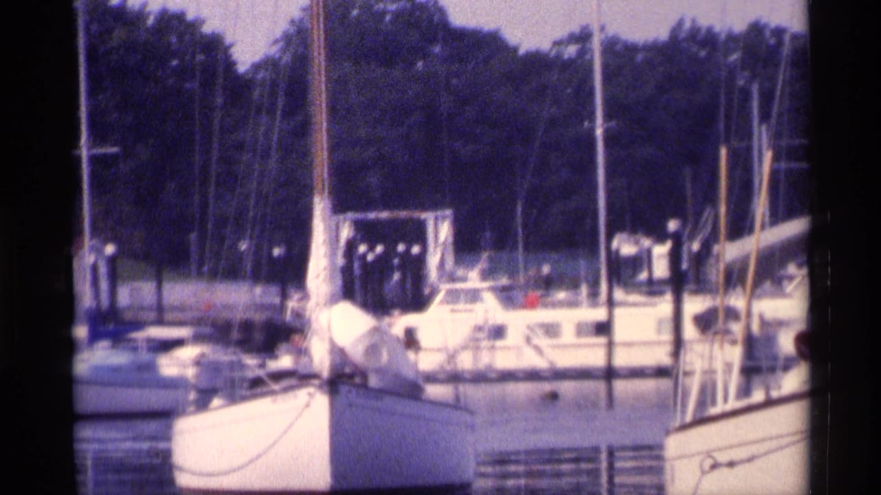 1973 New York marina view with white boats, tall masts, dock pilings, and a wooded shoreline seen through soft 8mm film color.