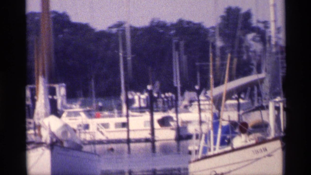 1973 New York 8mm frame showing white boats at a marina, vertical pilings, a wooded backdrop, and small distant people near the docks.