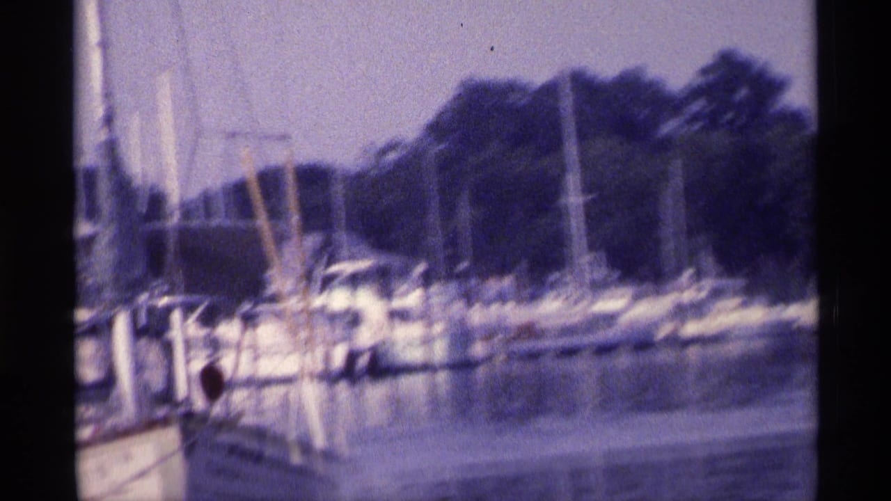 1973 New York home-movie frame with a slightly tilted view across marina water toward moored boats, sailboat masts, and dense trees.
