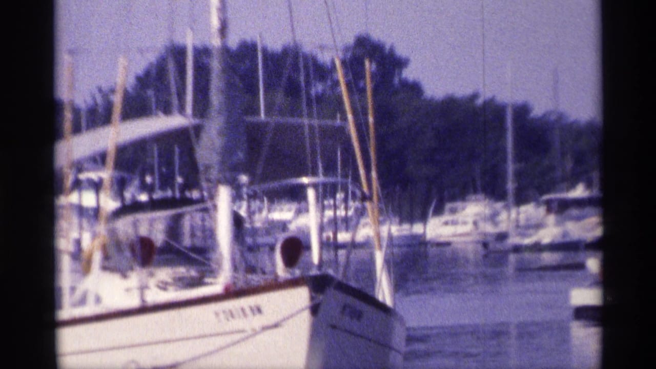Close 1973 New York marina frame showing a white sailboat bow, tall mast, dock posts, and other moored boats behind it.