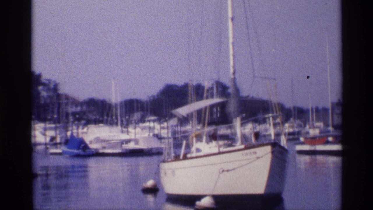 1973 New York marina shot with a white foreground sailboat, mooring buoys, clustered masts, and a low wooded shoreline.