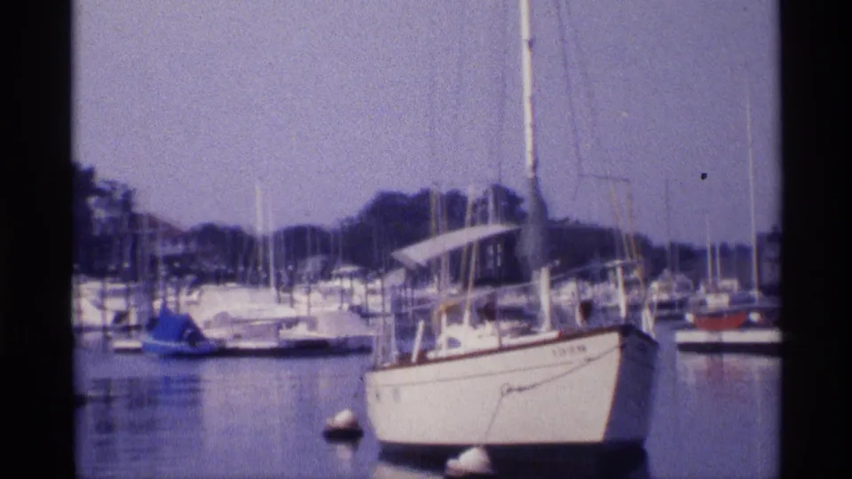 1973 New York marina shot with a white foreground sailboat, mooring buoys, clustered masts, and a low wooded shoreline.