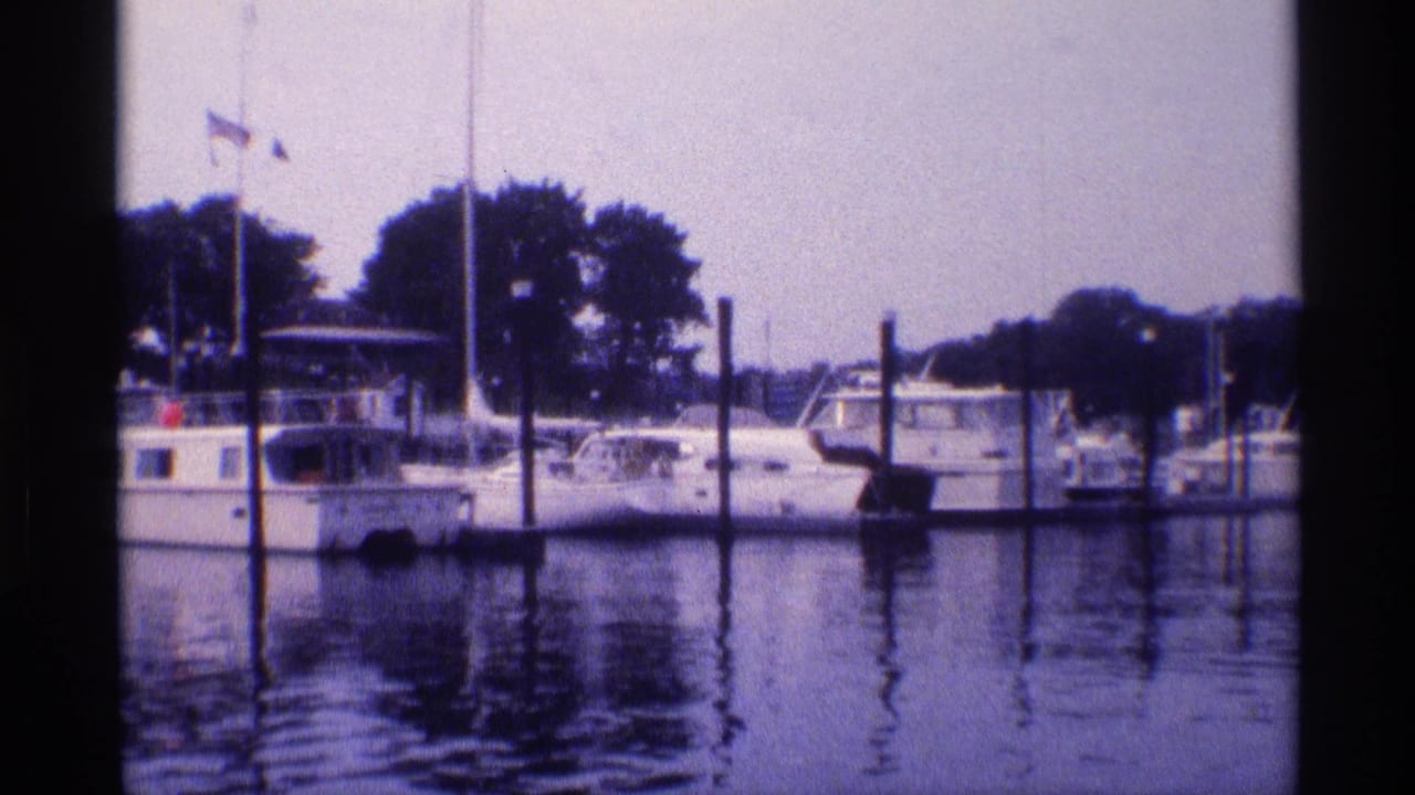 1973 New York 8mm frame showing docked white boats, marina pilings, small flags, and dark tree shapes along the shore.