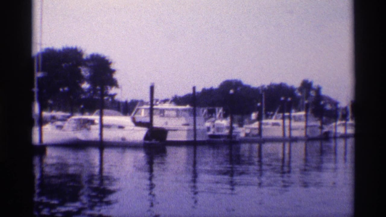 1973 New York home-movie view of moored sailboats and cabin boats behind marina pilings, with trees in the background.