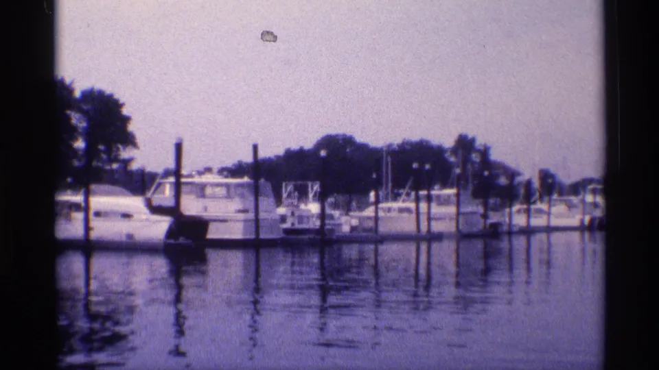 1973 New York 8mm frame showing white boats moored at a marina, with dock pilings, tree-lined shore, and water reflections.