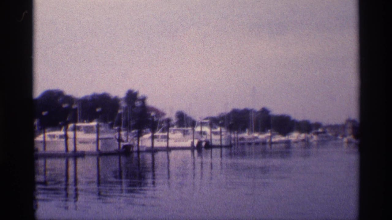 1973 New York 8mm frame of a quiet marina with white boats, dock pilings, rippling reflections, and a wooded shoreline.