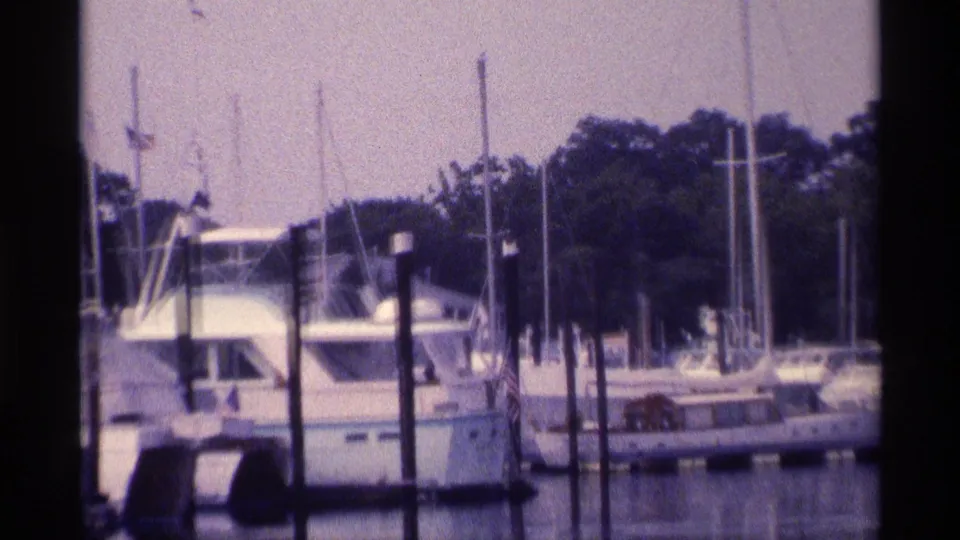 A soft 8mm close view shows a cabin boat beside dark dock pilings, with sailboat masts and a wooded shoreline behind.