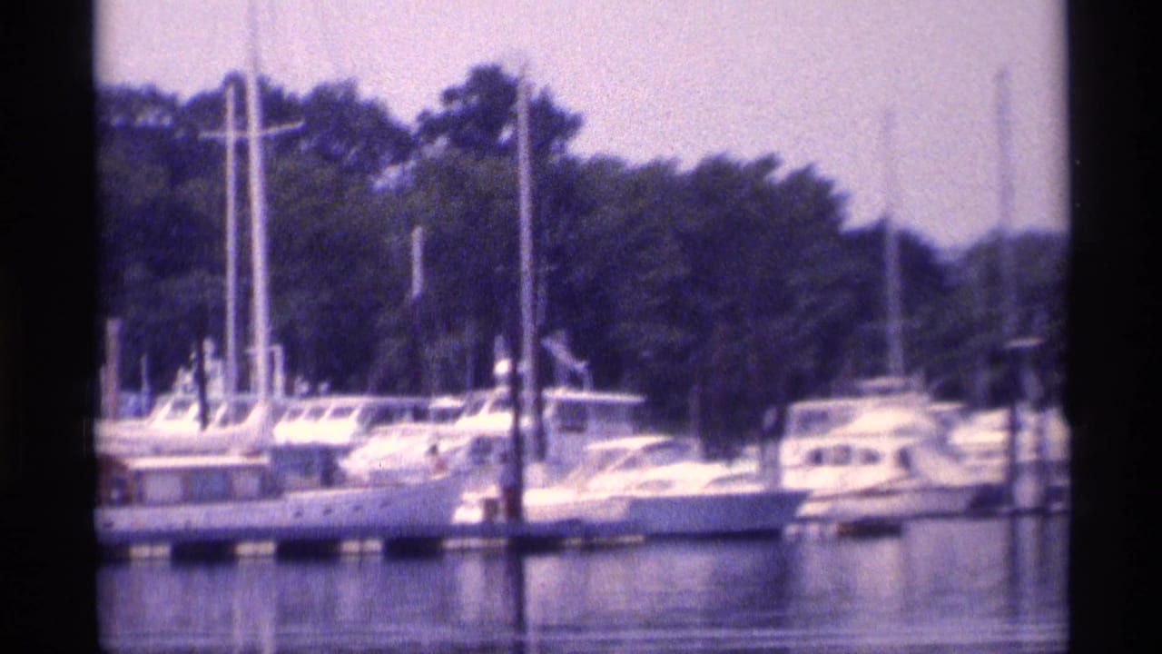 1973 New York marina view with moored sailboats, motor yachts, pilings, and a wooded shoreline in soft 8mm color.