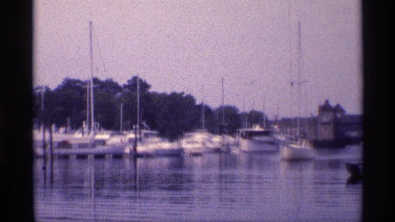 Wider 1973 New York 8mm view of a marina with docked boats, tall masts, calm water, trees, and a small waterside building.