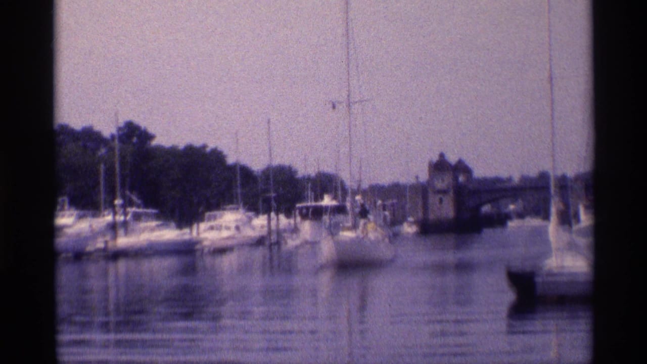 1973 New York 8mm frame of a small boat entering a marina channel with sailboats, pilings, trees, and a stone structure beyond.