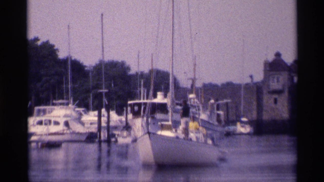 A closer 1973 New York marina frame shows sailboats, dock pilings, distant people on the dock, and a stone harbor structure.